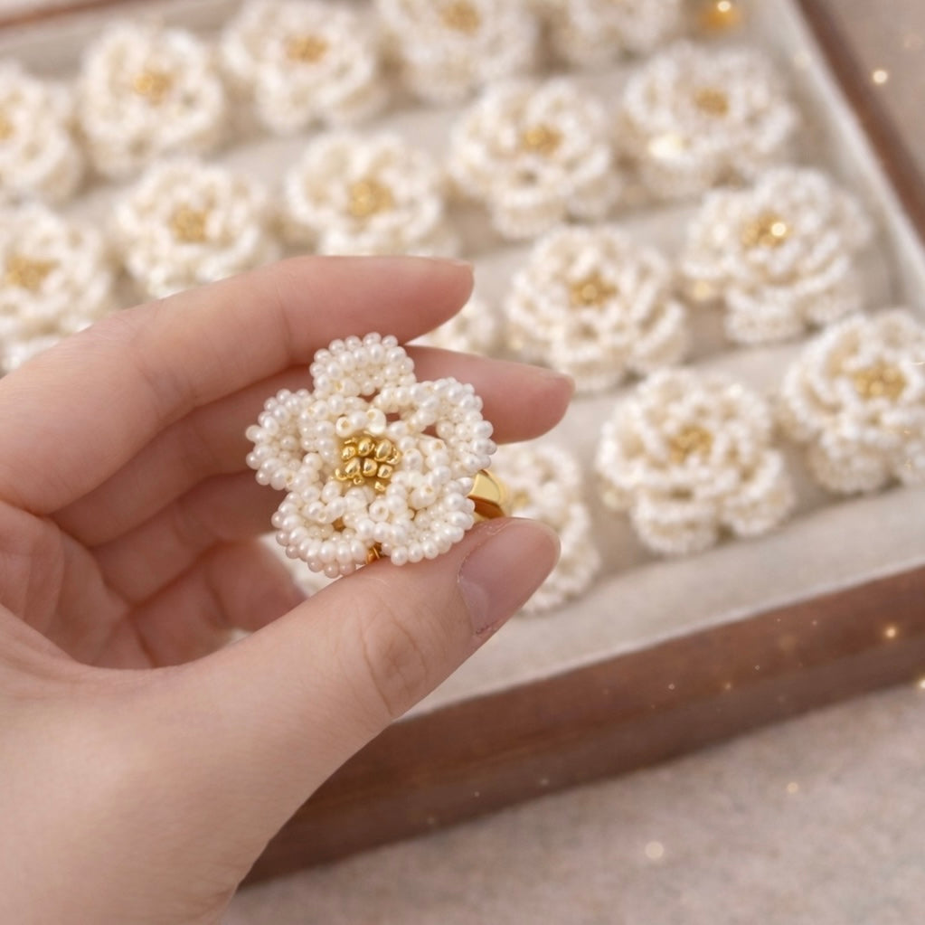 Hand holding a pearl flower ring with a tray of similar rings in the background.