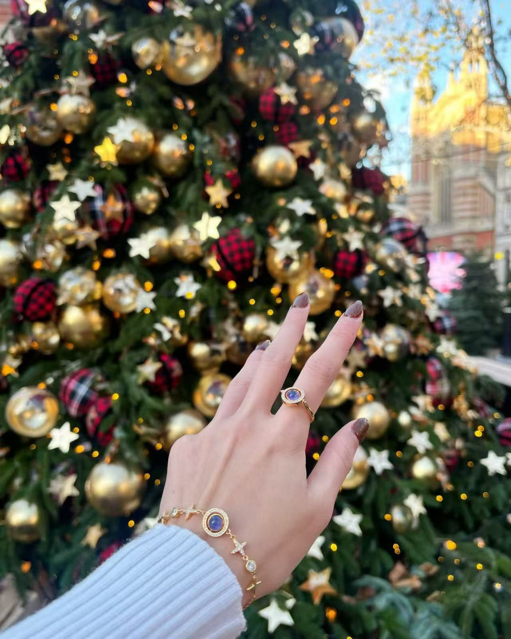 A festive outdoor scene featuring a hand wearing the Celestial Orbit Ring and Bracelet, both designed with gold star motifs and a transformable blue-violet gemstone. The jewelry is showcased in front of a Christmas tree decorated with golden ornaments, lights, and star accents, creating a warm and elegant holiday atmosphere.