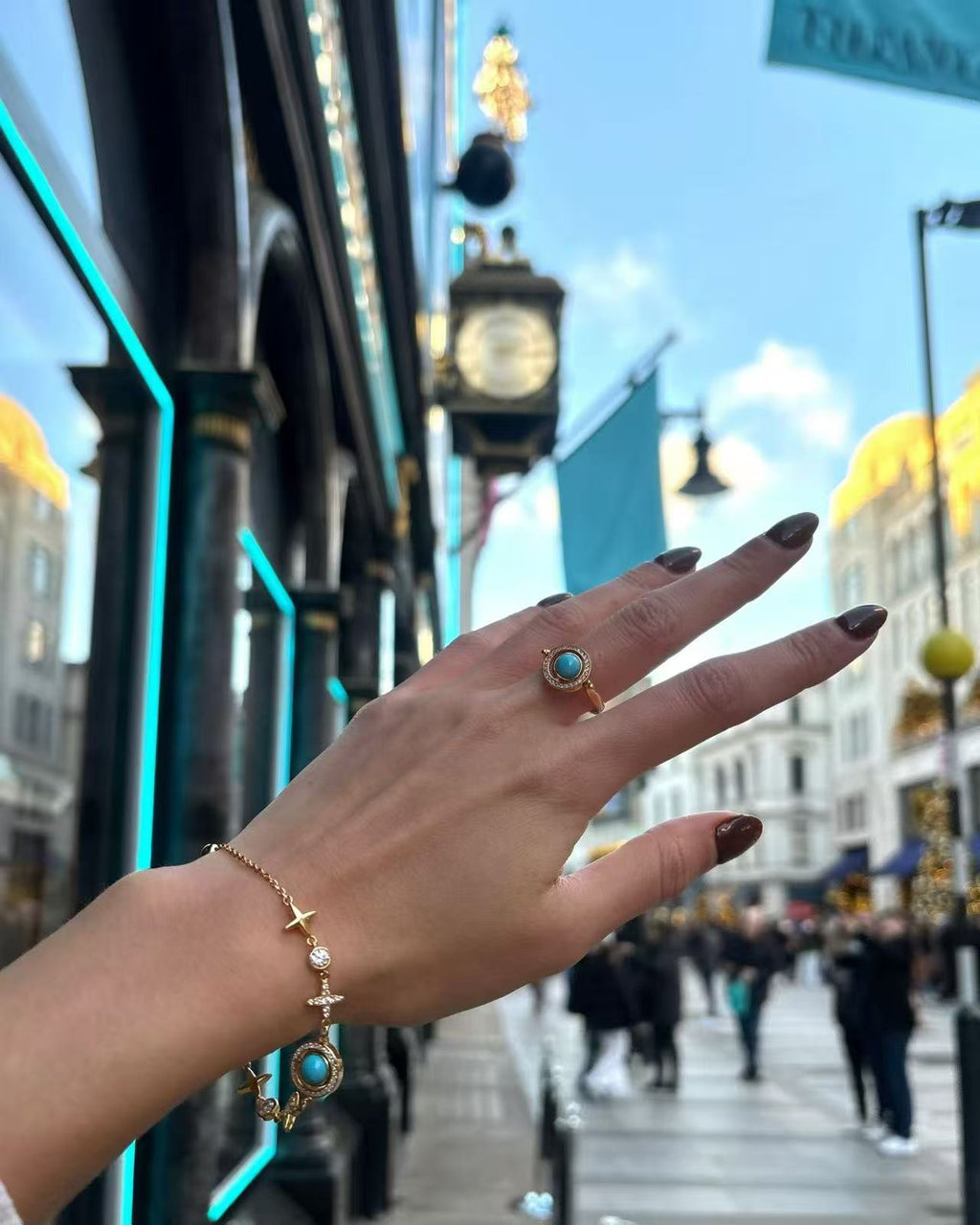 Hand wearing the Celestial Orbit Ring with turquoise-blue gemstones and gold star accents, photographed on a busy city street with reflective shop windows and pedestrians in the background.