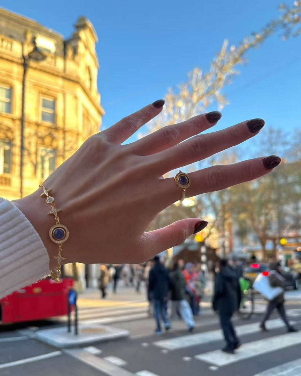 Hand wearing the Celestial Orbit Ring with blue-violet gemstones and gold star motifs, photographed outdoors above a busy pedestrian crossing with historic buildings in the background.