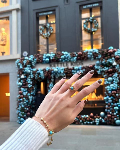 A hand wearing the Celestial Orbit Ring, featuring a transformable gemstone that shifts between blue and violet, set in a gold halo design; photographed outdoors with soft city architecture in the background.